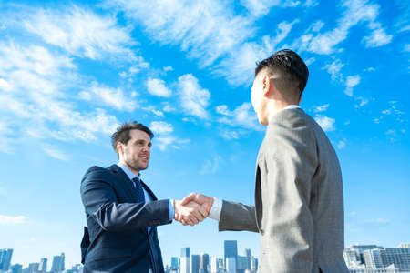 Businessmen shaking hands against the backdrop of the blue sky and buildings on fine dayの写真素材