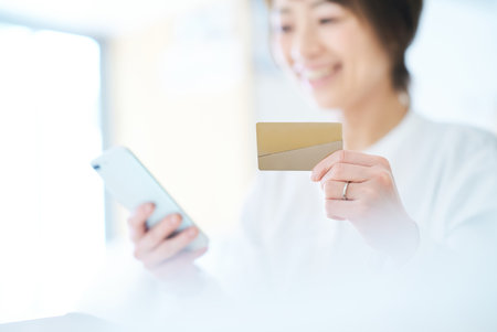 A woman holding a card and operating a smartphone in the roomの写真素材