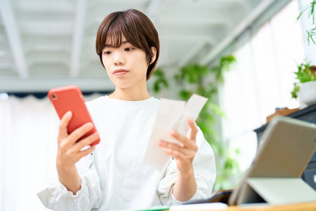 A woman holding a card and operating a smartphone in the roomの写真素材