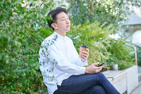 A man operating a smartphone while drinking coffee outdoorsの写真素材