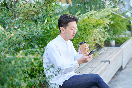 A man operating a smartphone while drinking coffee outdoorsの写真素材