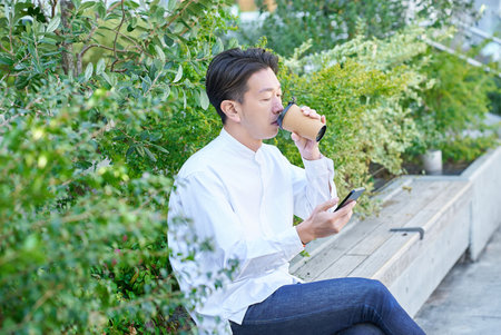 A man operating a smartphone while drinking coffee outdoorsの写真素材