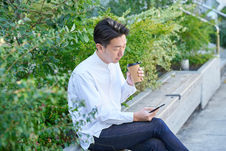 A man operating a smartphone while drinking coffee outdoorsの写真素材