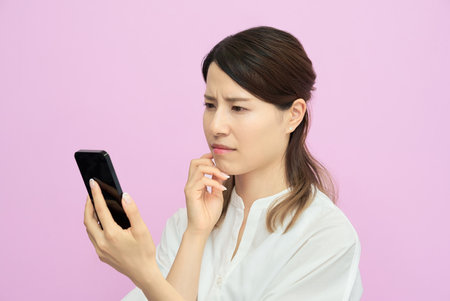 A woman looking at her smartphone screen with a troubled expression indoorsの写真素材