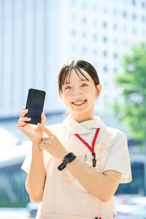 A young woman wearing a white coat and apron and holding a smartphone outdoorsの写真素材
