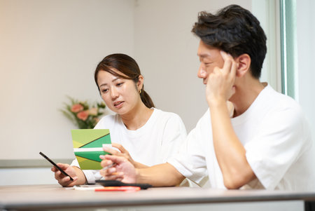 A man and a woman are thinking deeply while looking at their bank account passbook and smartphone in a roomの写真素材