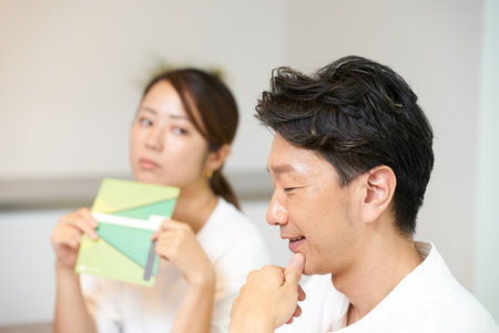 A man is looked at suspiciously by a woman holding a bankbook in the living roomの写真素材