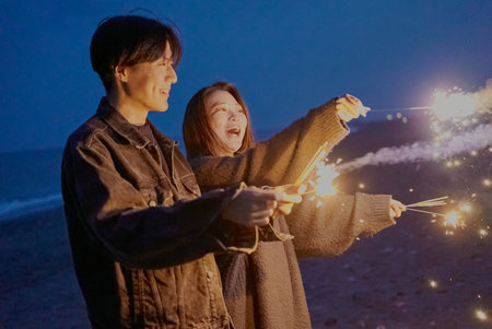 Young man and woman playing hand-held fireworks on the beach at nightの写真素材