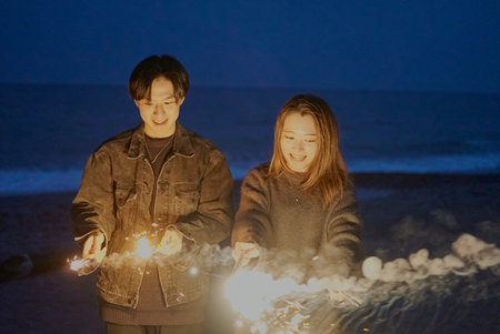 Young man and woman playing hand-held fireworks on the beach at nightの写真素材