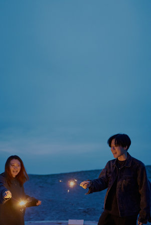 Young man and woman playing hand-held fireworks on the beach at nightの写真素材