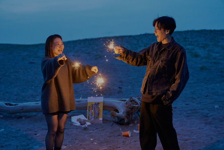 Young man and woman playing hand-held fireworks on the beach at nightの写真素材