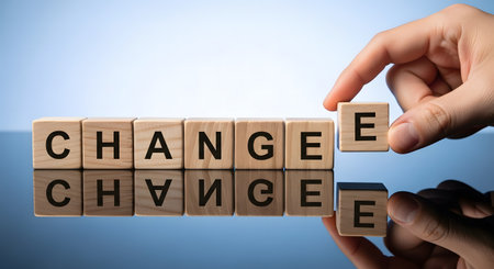 A hand is shown in the process of changing the word "CHANGE" by removing the last letter and placing it to form "CHANGEE". The letters are presented on individual wooden blocks, lined up and reflecting on a smooth surface. This visual represents the concept of transformation and the act of making a change.の素材