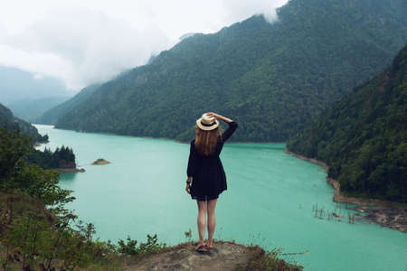 A girl in a hat and a dress stands on a stone on the background of the Ingur Reservoir in Svaneti....