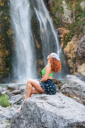 A girl in a green swimsuit sits under the streams of the waterfall. Grunas Waterfall is a picturesque site inside the National Park of Thethi, Albaniaの写真素材