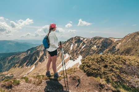 Girl hiker with a backpack stands on a rock in the mountains. Trekking life. Hike through the Carpathian mountains. Green mountain slopes and blooming rhododendron.の写真素材