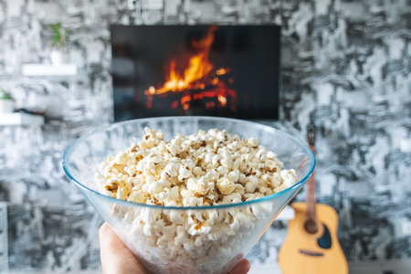 A glass bowl of popcorn and remote control in the background the TV works. Evening cozy watching a movie or TV series at homeの写真素材