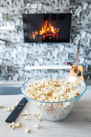 A glass bowl of popcorn and remote control in the background the TV works. Evening cozy watching a movie or TV series at homeの写真素材
