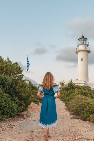 Girl in the blue dress standing on the hill next to white lighthouse, observing the sea below on the coast of Lefkada. Greeceの写真素材