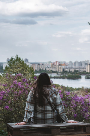 A girl in a grey shirt sits on a bench in the botanical garden of Kyiv during the lilac blossom season. Lilac is a symbol of Kyiv. Spring in the city.の写真素材