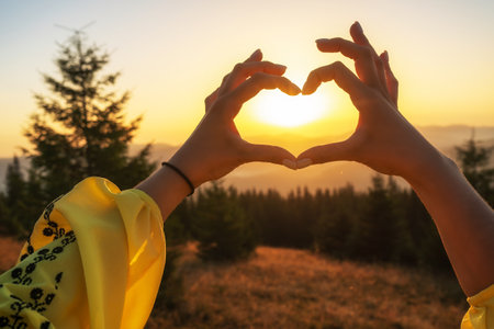 A girl holds her hands in the shape of a heart at dawn in the mountains. A girl in national Ukrainian clothes. Romance in the mountains.の写真素材