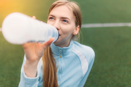 Girl after exercise, drinking water on the football field. Portrait of beautiful girl in sportswear. Isolated place for textの写真素材