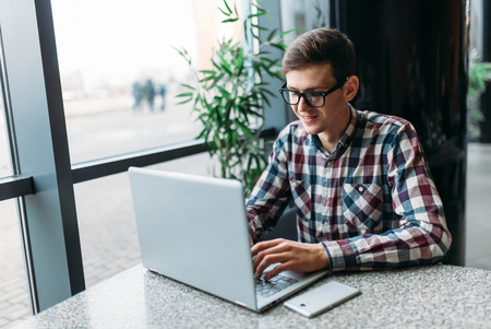 A man sits in a cafe and working on laptop, looking for work, the student writes the homework, a guy in a shirt and glasses, nerdの写真素材