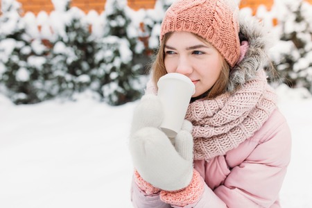Portrait of a beautiful young girl in white knitted mittens, outdoors, drinking a hot beverage, bright winter day.woman smiling and happy, macro, trees, first snow.beautiful imageの写真素材