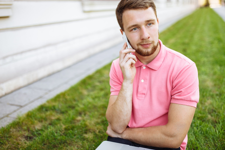 Handsome man sitting on the grass in the city with a laptop and talking on the phone, job search, rest on the Internetの写真素材