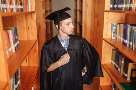 Successful graduate guy, in academic dresses, standing in the library, choosing books, can be used for advertising,の写真素材