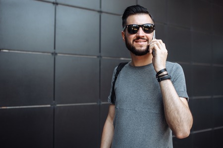Portrait of a beautiful stylish guy, hipster with glasses, talking on the phone, dressed in a gray empty t-shirt, standing on a black wall background.の写真素材