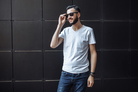 Photo of a beautiful stylish guy, a hipster with glasses, dressed in a white empty t-shirt, standing on a black wall background. .の写真素材