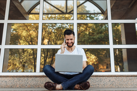Portrait of a beautiful stylish guy wearing a white t-shirt, who sits on a parapet, works with a laptop and talks on the phone, on the street in front of a glass building,の写真素材
