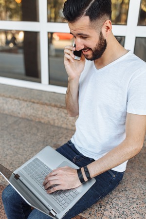 Portrait of a beautiful stylish guy wearing a white t-shirt, who sits on a parapet, works with a laptop and talks on the phone, on the street in front of a glass building,の写真素材