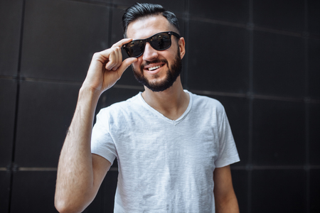 Photo of a stylish guy, a hipster with glasses, dressed in a white empty t-shirt, standing on a black wall background.の写真素材