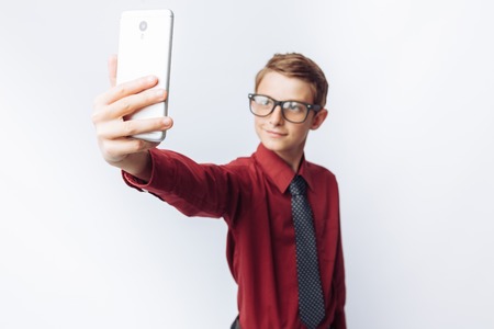 Portrait of a positive and emotional schoolboy, takes a selfie on your smartphone, white background, glasses, red shirt, business theme,の写真素材