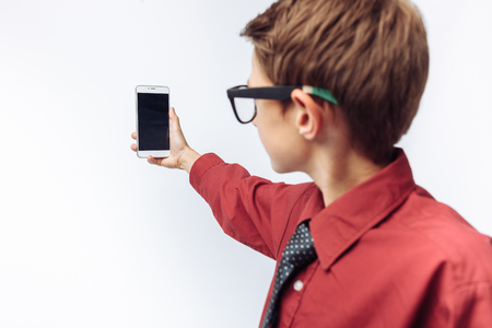 Portrait of a positive and emotional schoolboy, takes a selfie on your smartphone, white background, glasses, red shirt, business theme,の写真素材