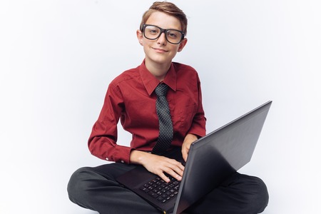 Portrait of positive and emotional schoolboy posing with laptop, typing on keyboard, white background, glasses, red shirt, business theme,の写真素材