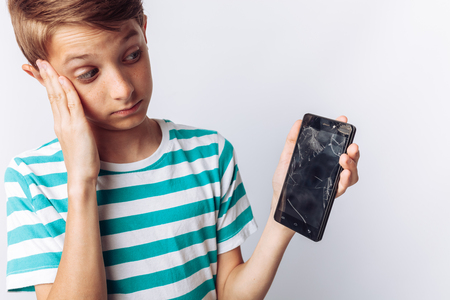 Portrait of a beautiful and emotional boy, in whose hands a broken phone shows surprise, white background, blue t-shirt,の写真素材
