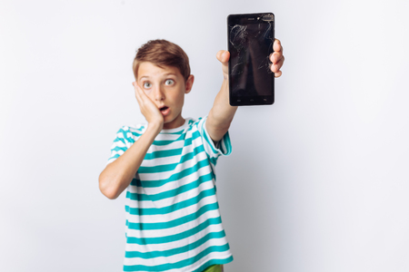 Portrait of a beautiful and emotional boy, in whose hands a broken phone shows surprise, white background, blue t-shirt,の写真素材