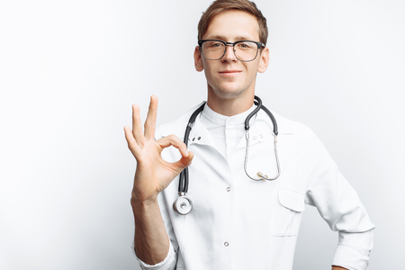 Portrait of a young doctor, on a white background, which shows the gesture Okey, trainee in the Studio, with a stethoscope on the neck,の写真素材