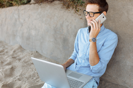 Young guy with glasses, working on his laptop on the beach against the gray wall, at sunset, working on vacation,の写真素材