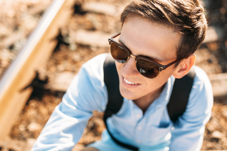 Beautiful stylish guy with glasses, posing sitting on the rails of the train at sunset, hipster posing in stylish clothes,の写真素材