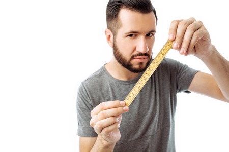 Handsome young hipster guy, in a grey t-shirt, holds a centimeter in front of his face and looks at him, on a white backgroundの写真素材
