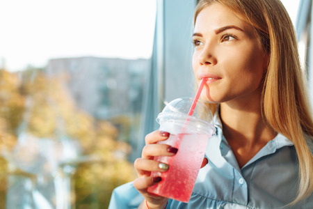 Portrait of a young and beautiful girl standing near the window in the room with a cocktail in her handsの写真素材