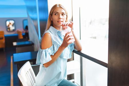 Beautiful young cheerful girl, drinking a cocktail, sitting in a cafe and waiting for someoneの写真素材