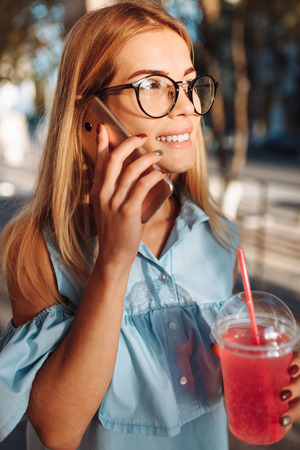 Young beautiful girl student, wearing glasses, talking on the phone in a good mood, and holding a cocktailの写真素材