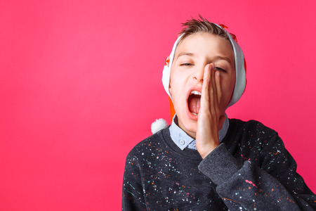 Positive teen, in Santas hat, shouts "merry Christmas" isolated on red backgroundの写真素材