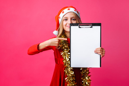 girl in Santa hat and tinsel on the neck, holds the tablet with a clean sheet and pointing a finger to plan for the New year on red backgroundの写真素材