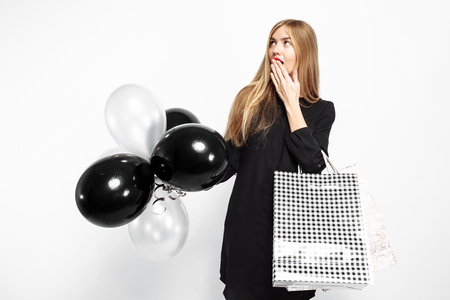 Elegant shocked girl, in black dress, with shopping bags and black balls in her hands isolated on white background . Black Friday, shopping, discountsの写真素材