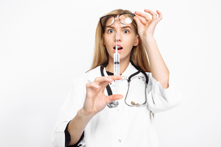 Young scared intern doctor wearing glasses with a stethoscope around his neck, holding a syringe in his hands and looking at him with fear, on a white backgroundの写真素材